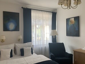 Ett hotellrum med en säng, två vägglampor, en stol, ett skrivbord och två tavlor.