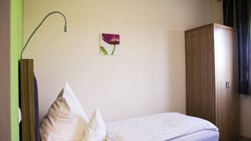 A neatly made bed with white bedding, a single wall-mounted lamp, a wooden wardrobe, and a small framed picture of a flower on the wall.
