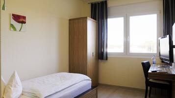 A bedroom with a bed, a wooden wardrobe, a desk with a chair, a television, and a window with curtains.