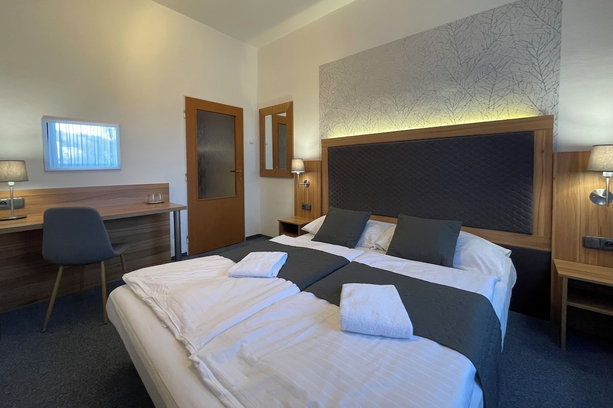 A hotel room with a large bed, a desk, a chair, a mirror, and a wall-mounted lamp.