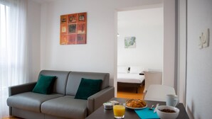 Una habitación de hotel moderna con un sofá gris, una pequeña mesa de comedor con alimentos para el desayuno, una cama y una silla blanca.