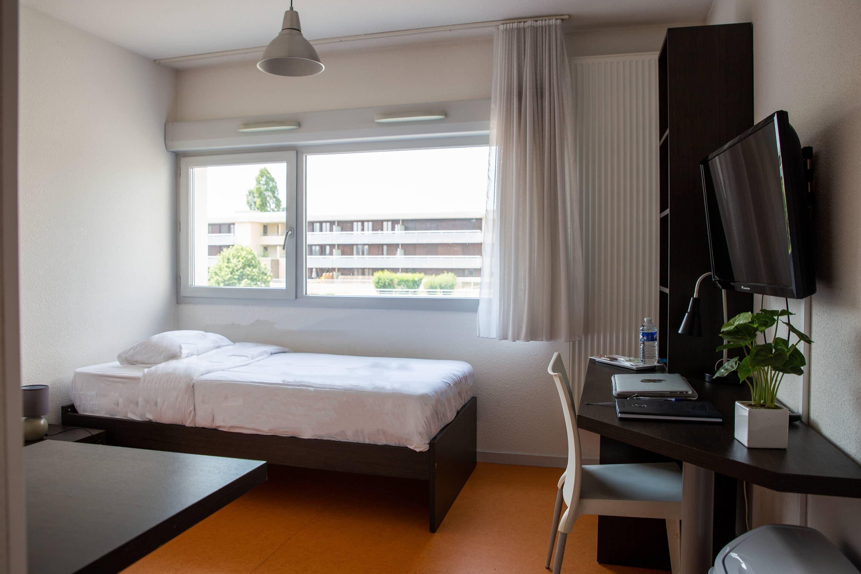 Habitación de hotel con cama, escritorio, silla, televisor y ventana con vista a edificios y árboles.