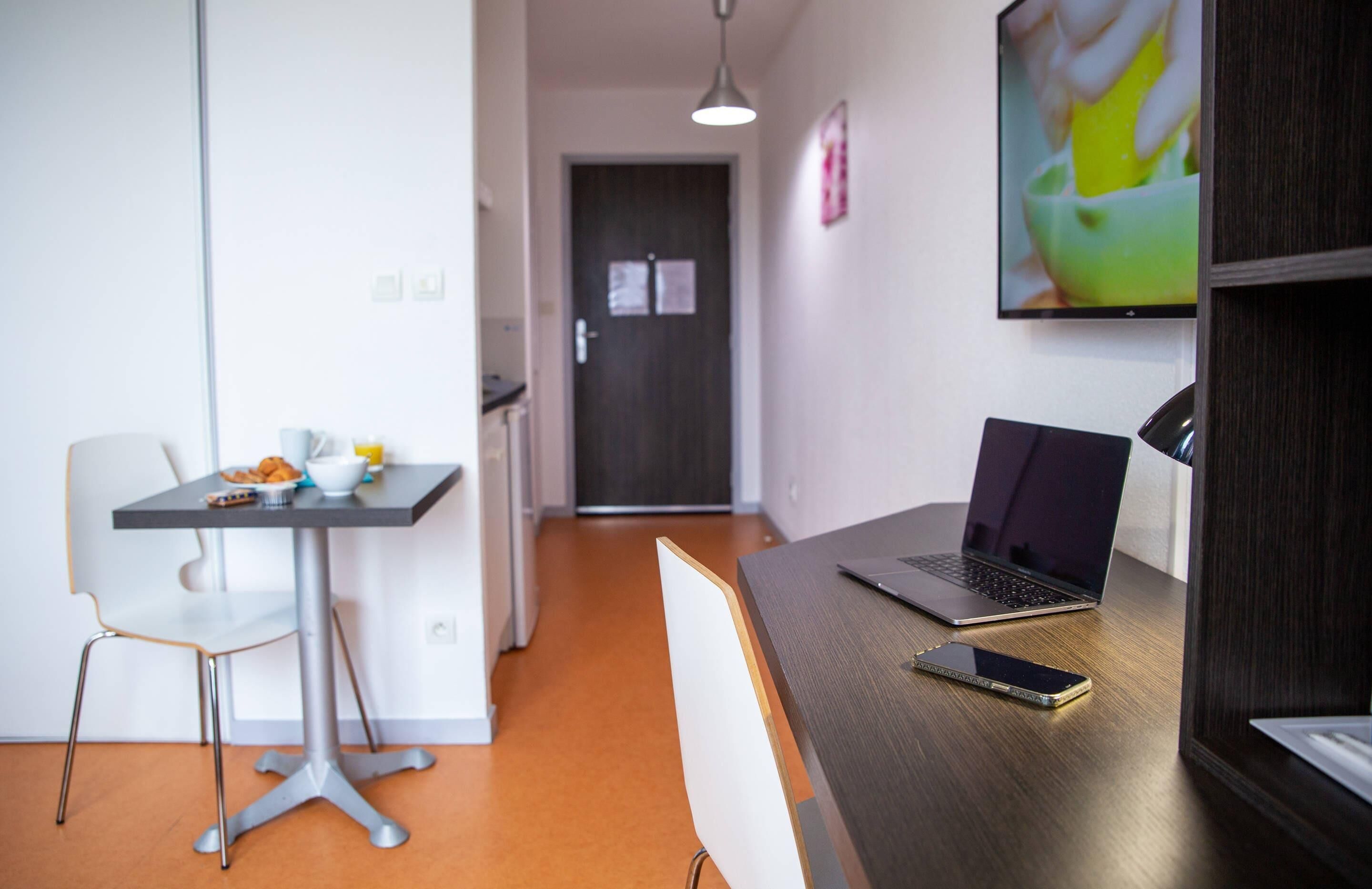 Un espacio de oficina moderno con un escritorio, laptop, teléfono inteligente y una silla. Se observa una puerta al fondo y un televisor montado en la pared.