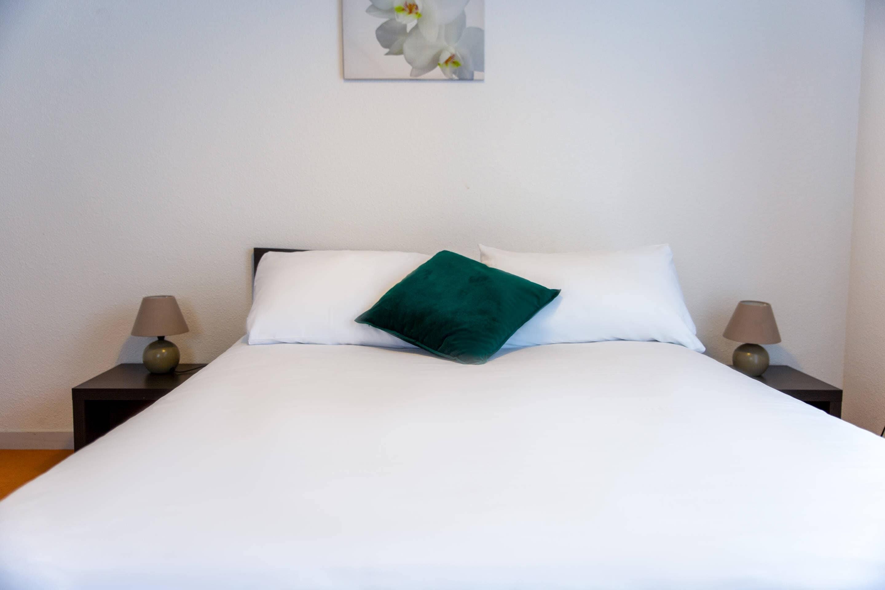 A neatly made bed with white linens and a single green pillow, flanked by two bedside tables with lamps.