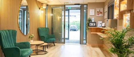 A reception area with a wooden wall, green chairs, a small table, and a reception desk with a plant.