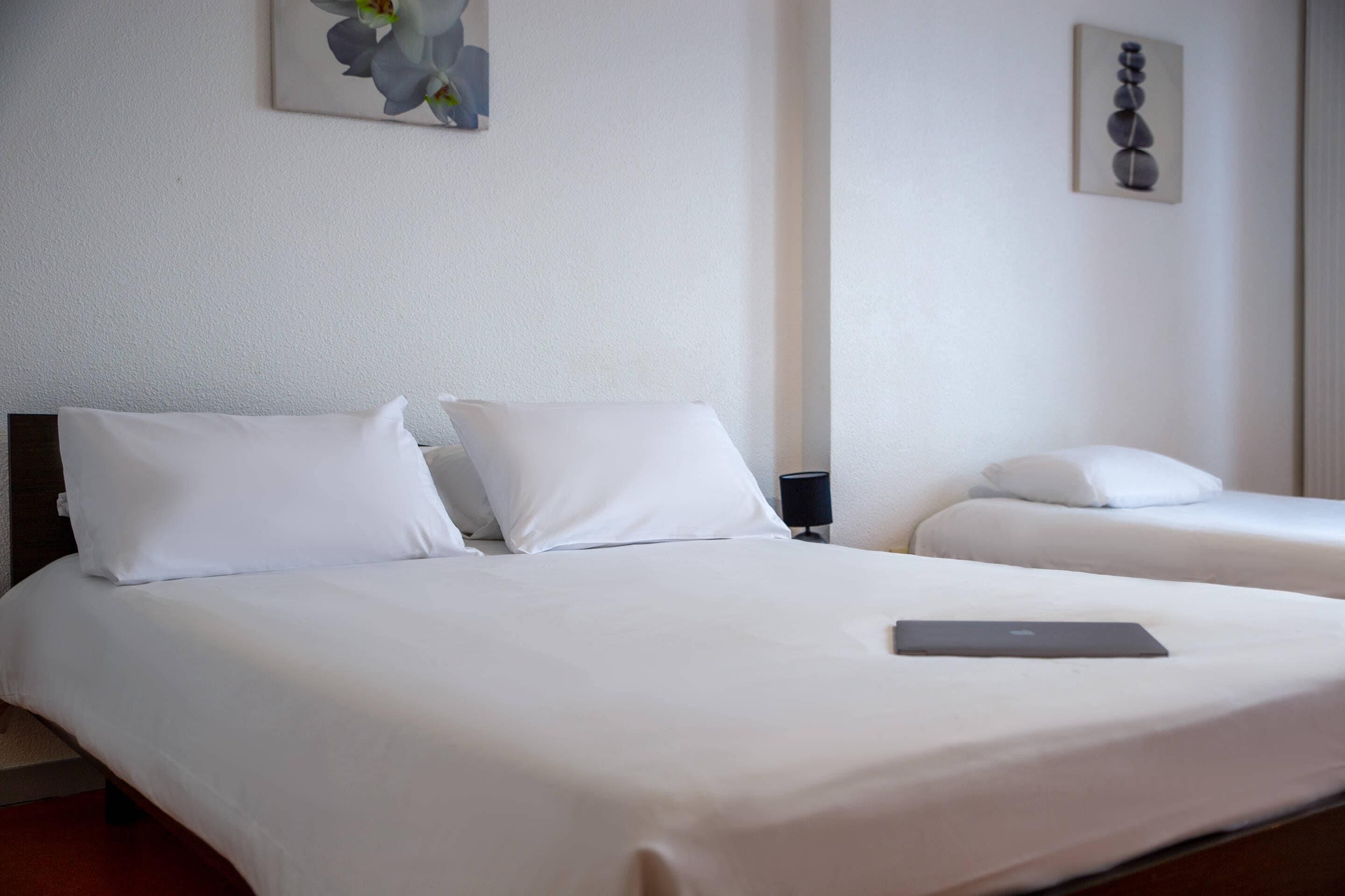 Habitación de hotel con dos camas, ropa blanca, un ordenador portátil sobre una cama y obras de arte en las paredes.