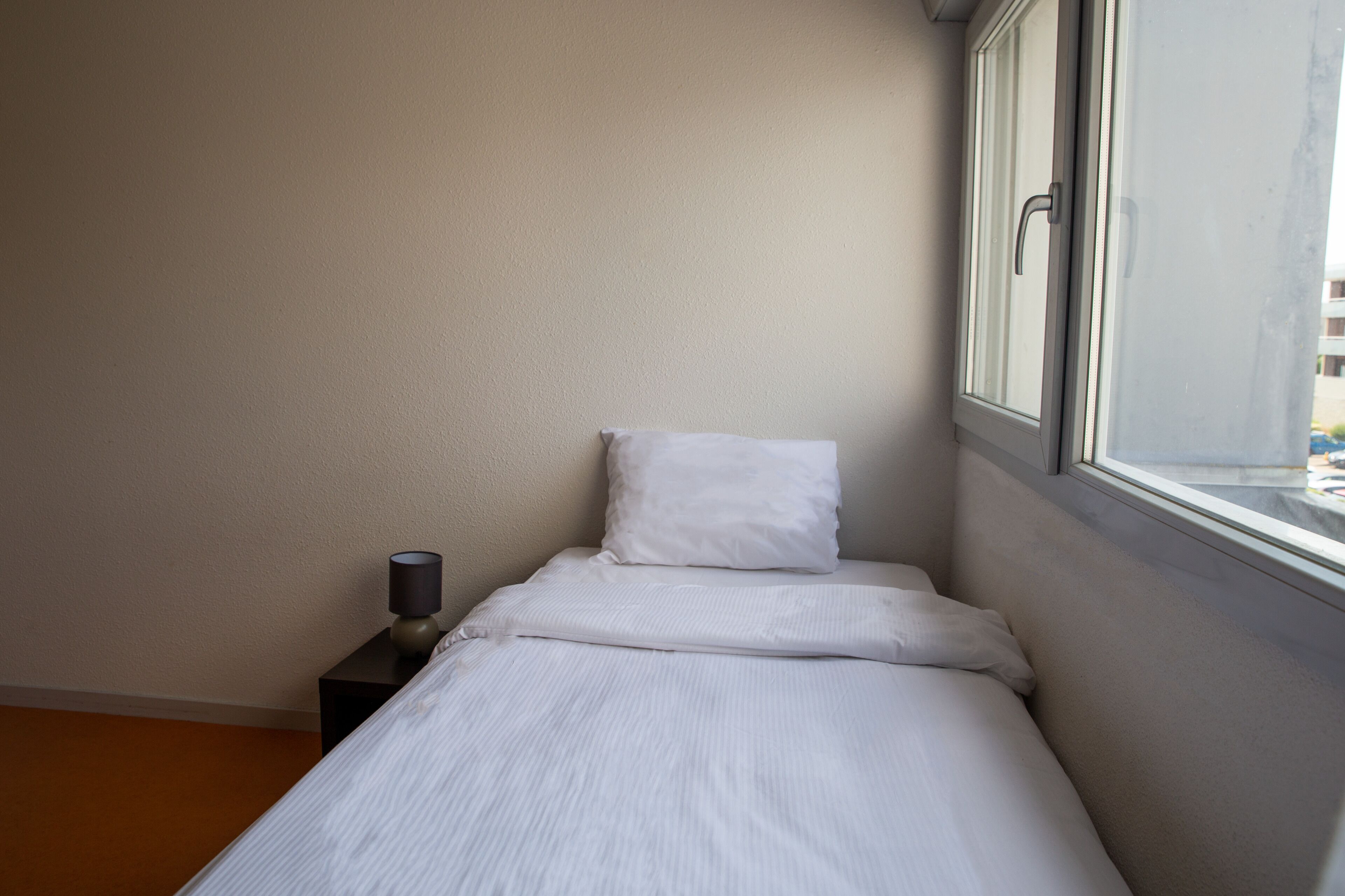 Ein kleines, ordentlich bezogenes Bett mit weißen Laken in einem Zimmer mit Fenstereck.