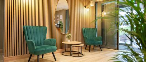 Un área de espera moderna con dos sillones verdes, una pequeña mesa redonda y un espejo en la pared.