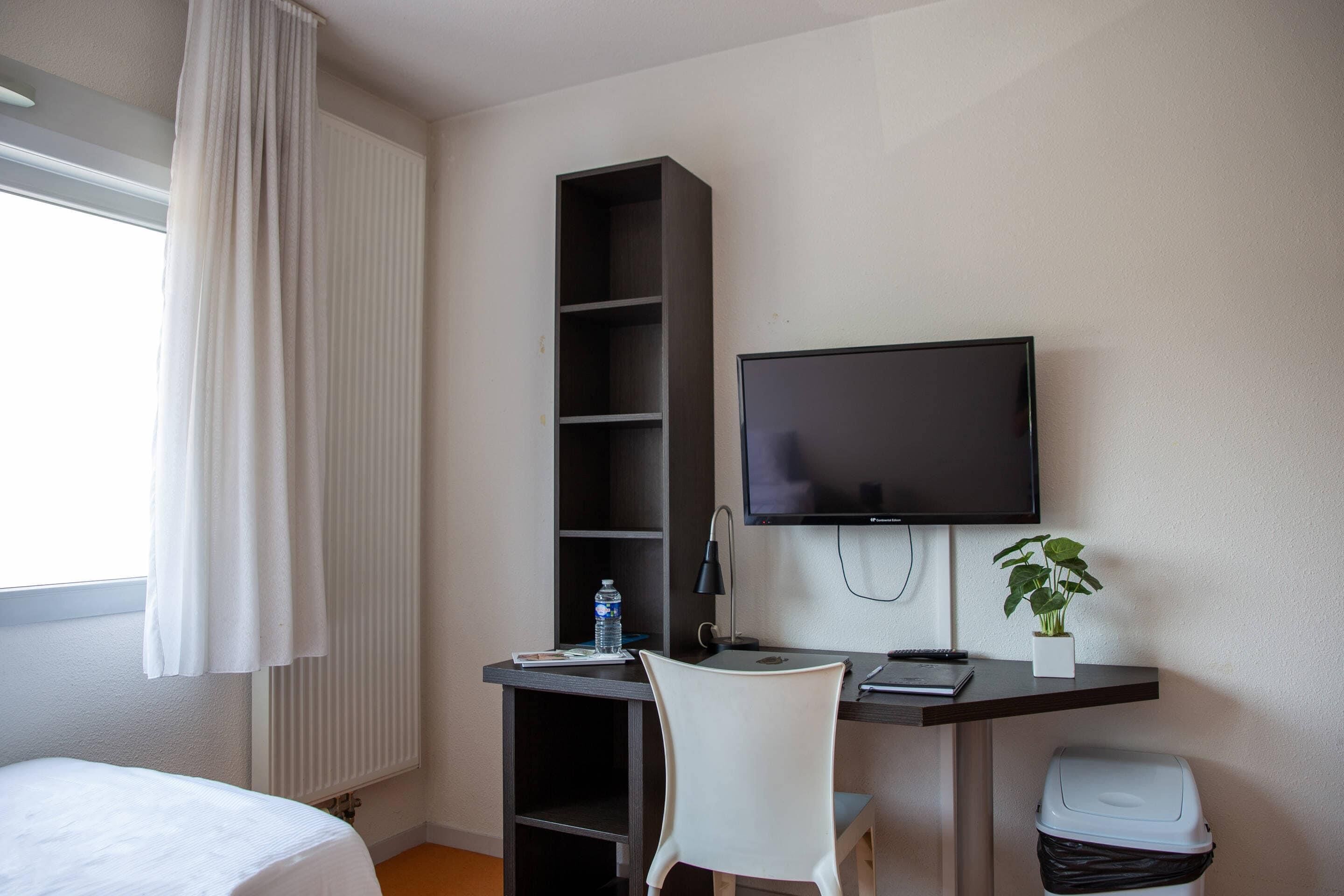 Ein Hotelzimmer mit einem Bett, einem Schreibtisch mit Computer, einem Fernseher, einem Bücherregal, einer Pflanze und einem Mülleimer.