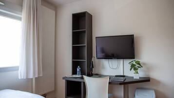 A hotel room with a bed, a desk with a computer, a TV, a bookshelf, a plant, and a trash bin.
