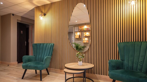 Una habitación con dos sillones verdes, una mesa redonda de madera y un espejo grande en una pared de listones de madera.
