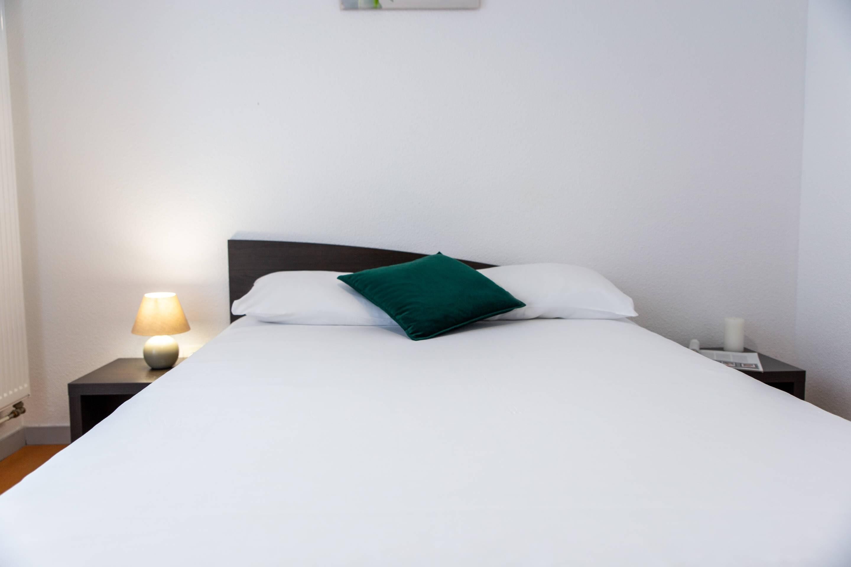 Una cama bien hecha con sábanas blancas y un almohadón verde, junto a una lámpara de noche y una mesita con una vela.