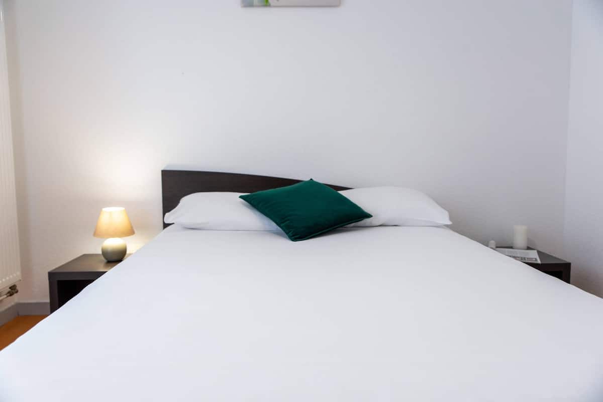 Una cama bien hecha con sábanas blancas y un almohadón verde, junto a una lámpara de noche y una mesita con una vela.