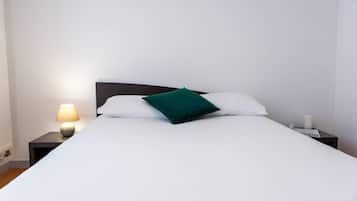 Ein ordentlich bezogenes Bett mit weißen Laken und einem grünen Kopfkissen, dazu eine Nachttischlampe und ein kleiner Tisch mit einer Kerze.