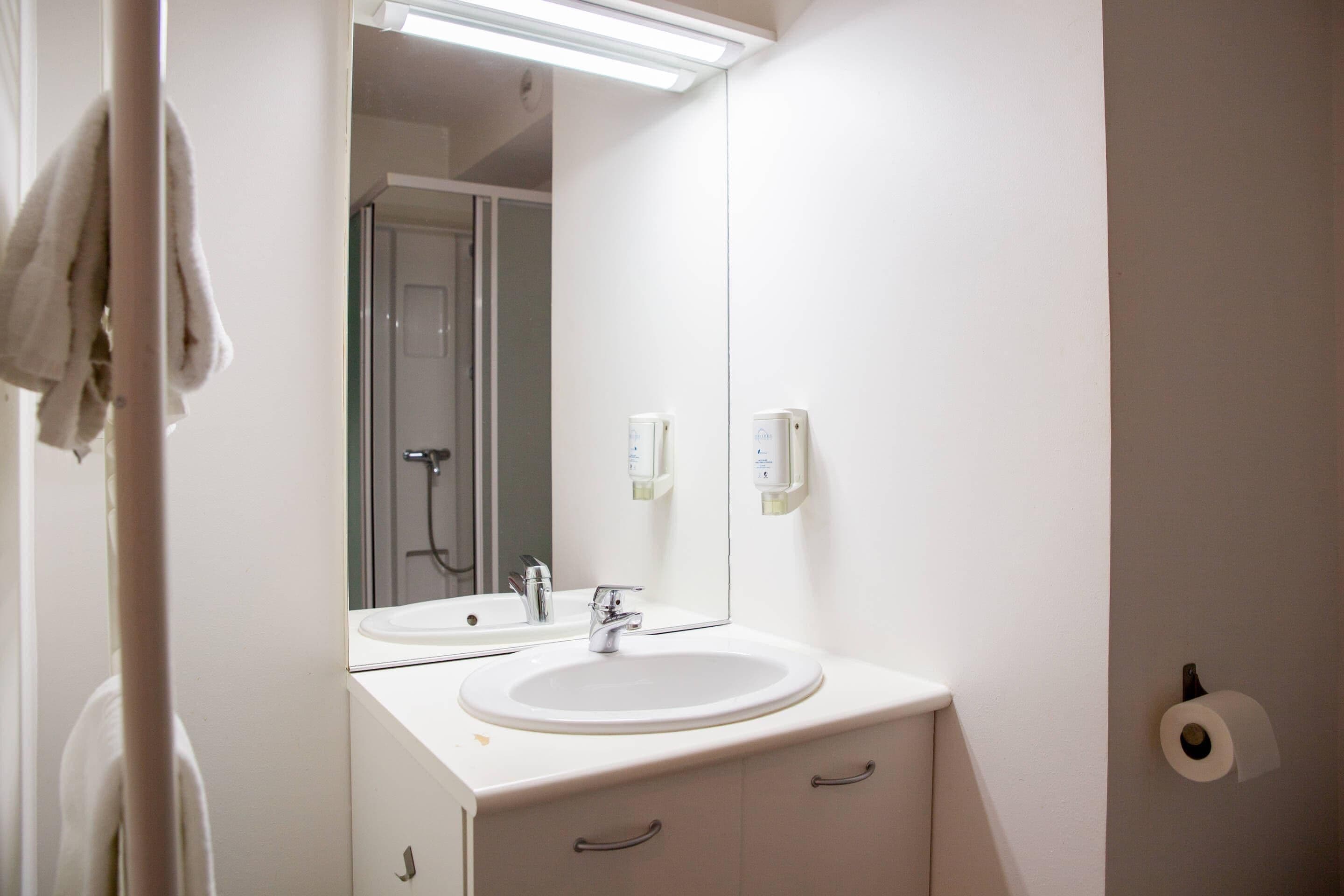 Ein Badezimmer mit einem weißen Waschbecken, einem Spiegel, einem Handtuch, das an einem Halter hängt, und einem Toilettenpapierhalter an der Wand.