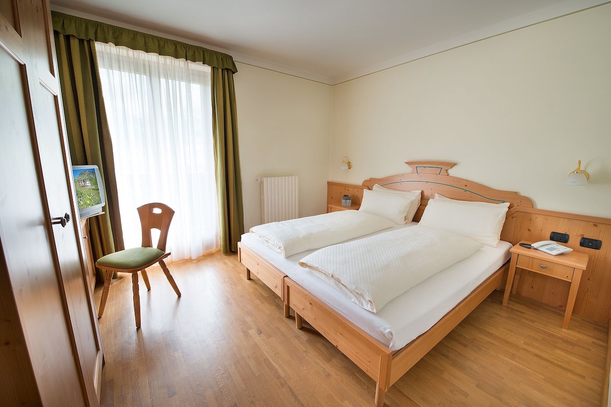 Un dormitorio con una cama de madera, una mesita de noche con un teléfono, una silla y una ventana con cortinas.