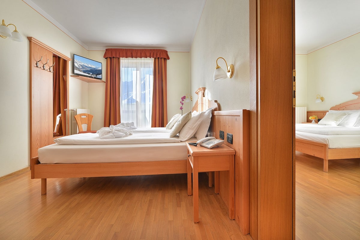 Habitación de hotel con una cama de madera, una mesita de noche con teléfono, una ventana con cortinas y un televisor montado en la pared.