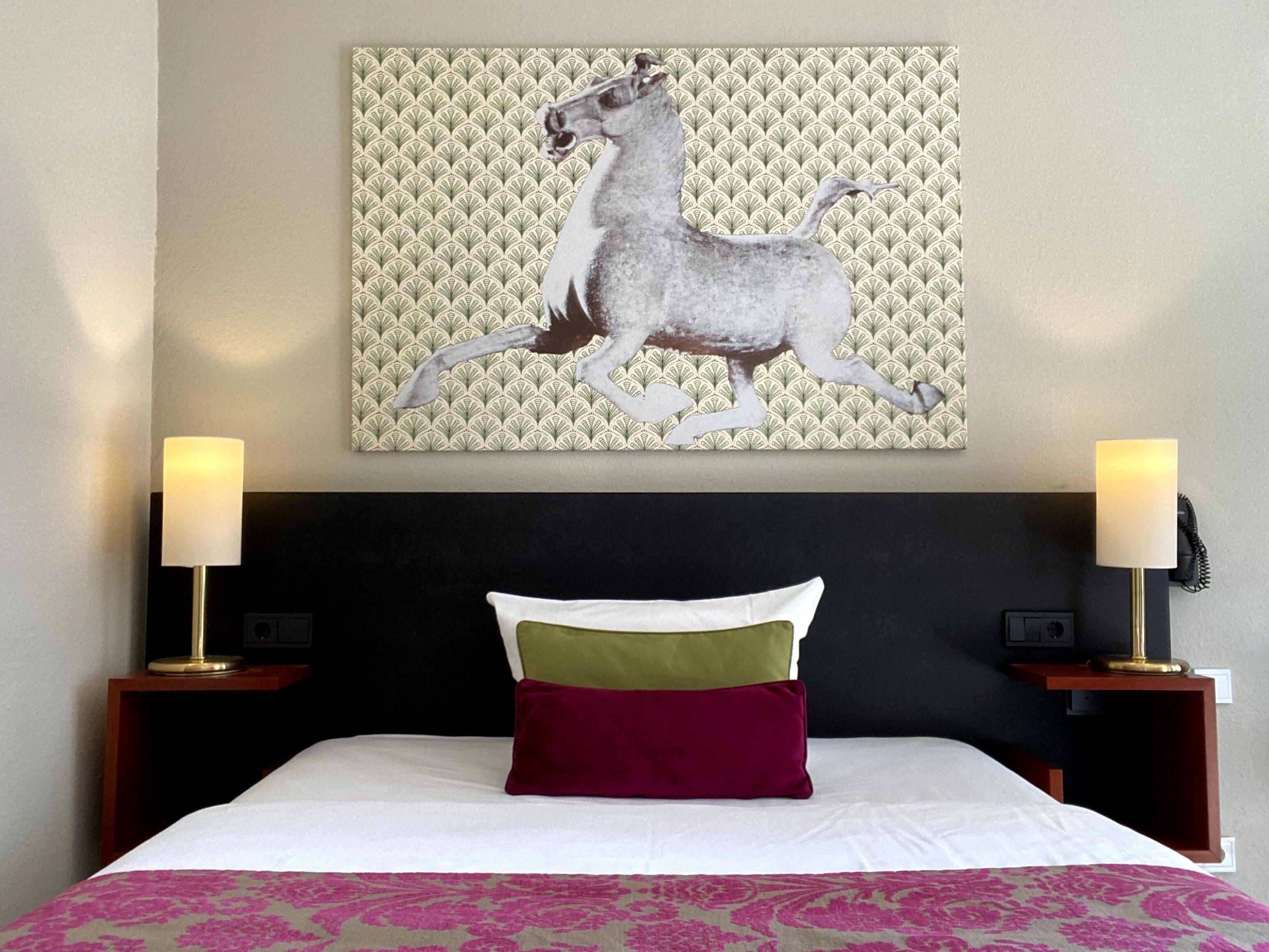 A hotel room with a bed, two bedside lamps, a headboard, and a wall-mounted artwork of a horse.