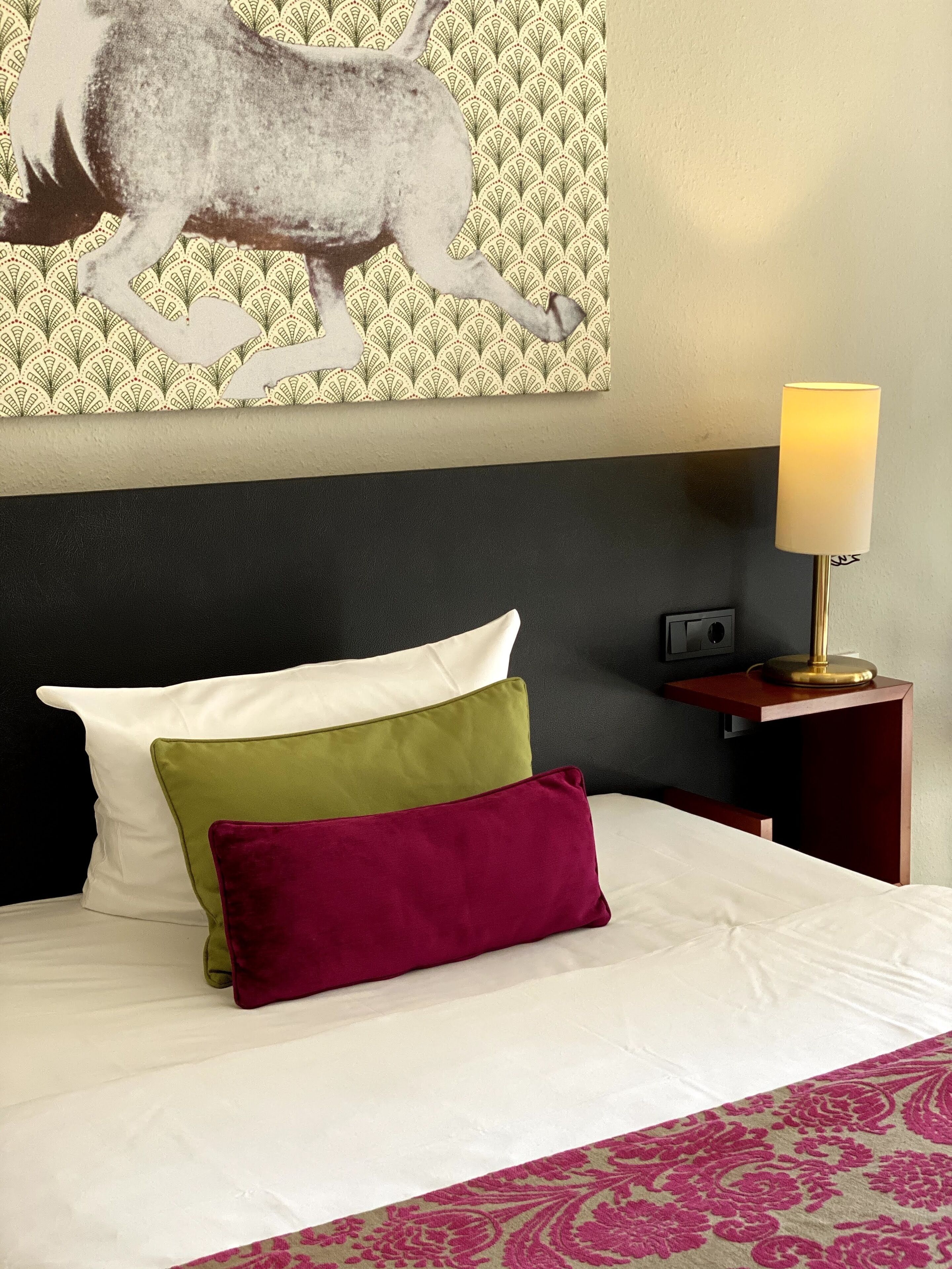 Ein Hotelzimmer mit Bett, Kissen, Nachttisch, Lampe und Wandbild.