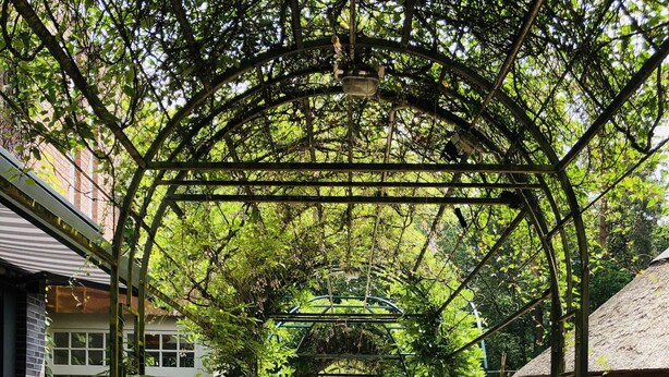 A covered pathway lined with tables and chairs, surrounded by greenery and a brick building. - Heidegrund (Garrel)