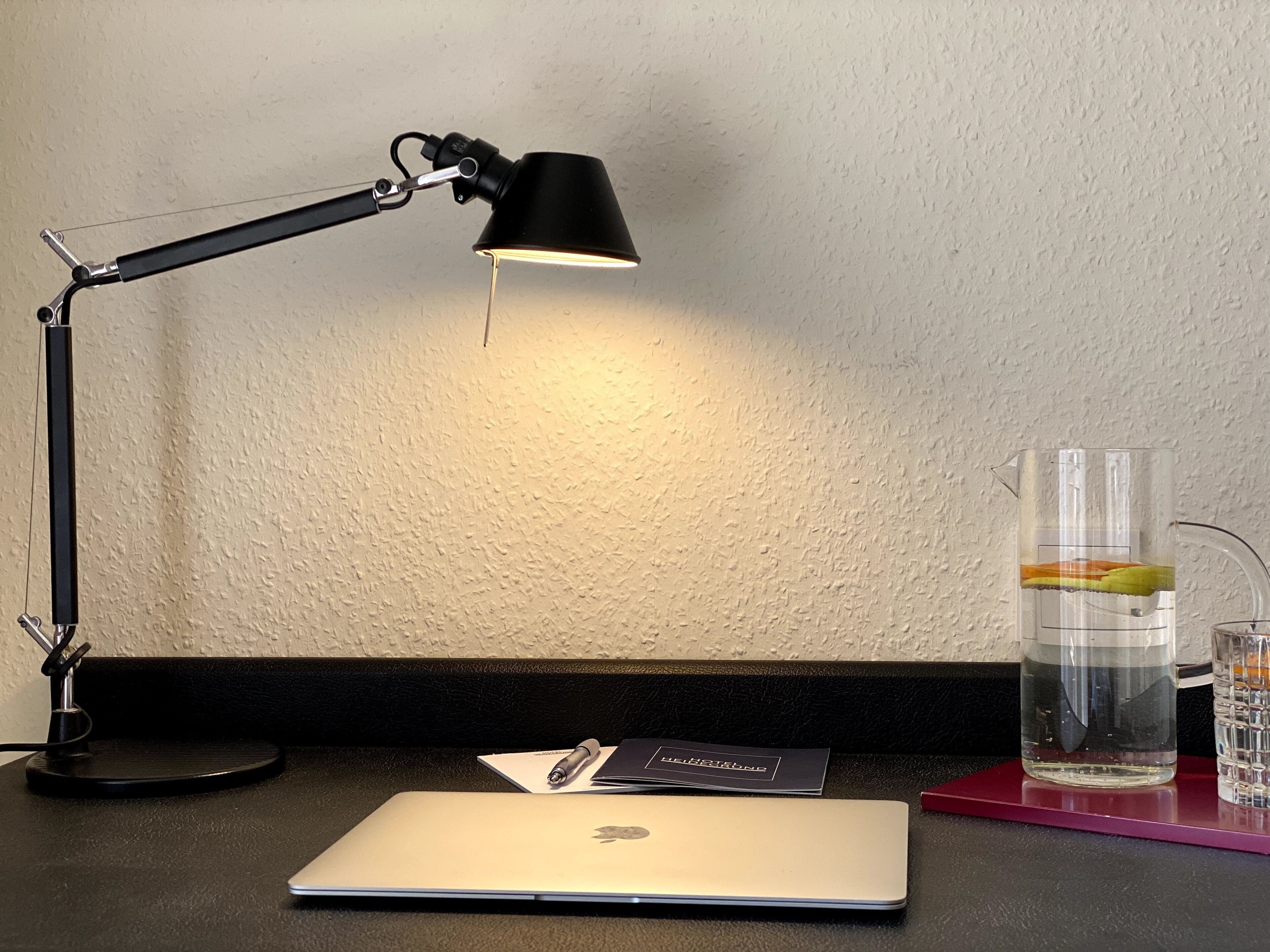 Un escritorio con una computadora portátil, un bolígrafo, una libreta, un vaso de agua y una lámpara.