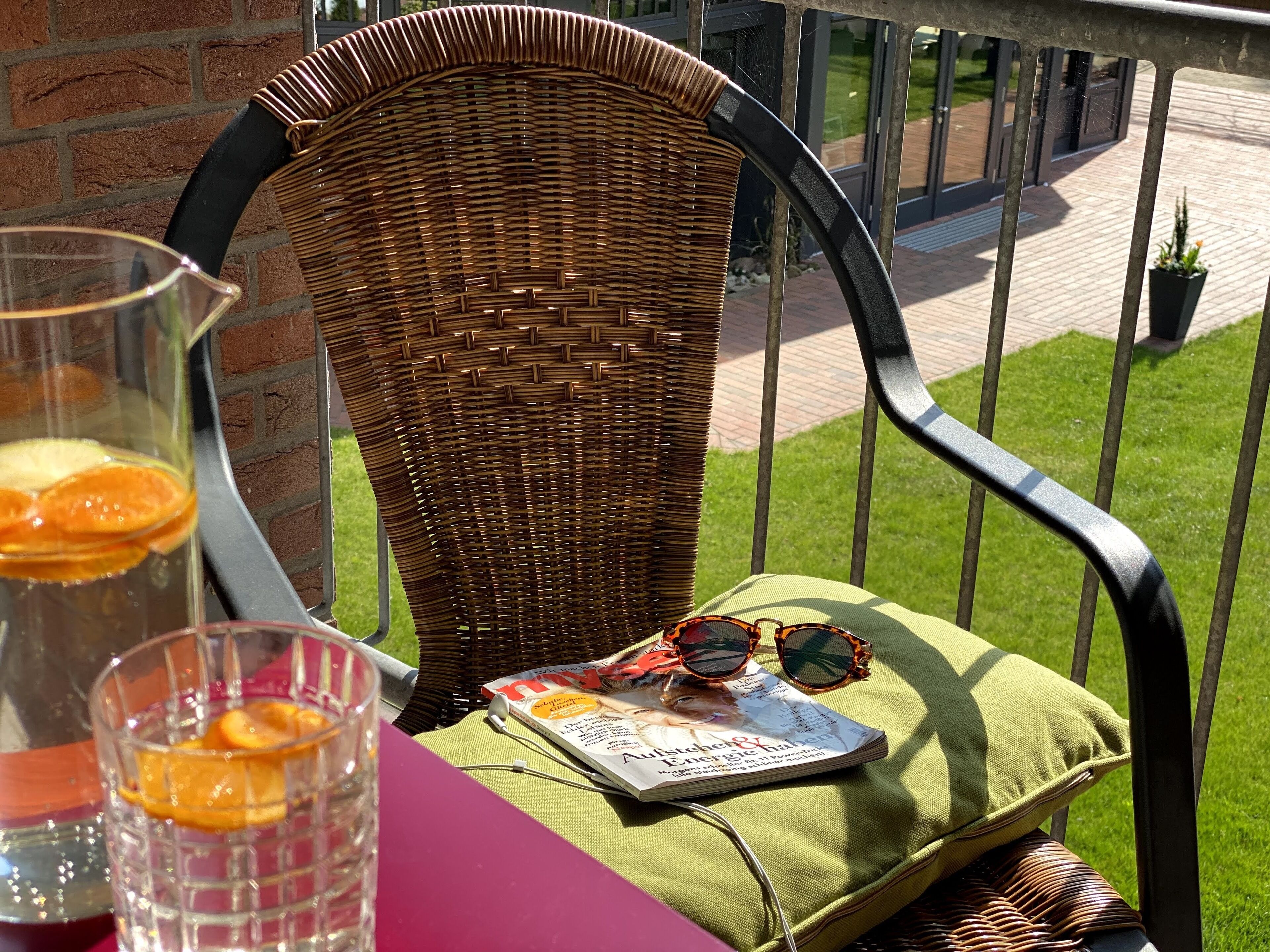 A wicker chair with a cushion, a glass of orange slices, and a magazine on a table outdoors.