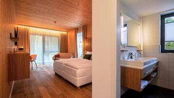 Een moderne hotelkamer met een houten plafond, een groot bed, een zithoek met een fauteuil, en een badkamer met een grote spiegel en wastafel.