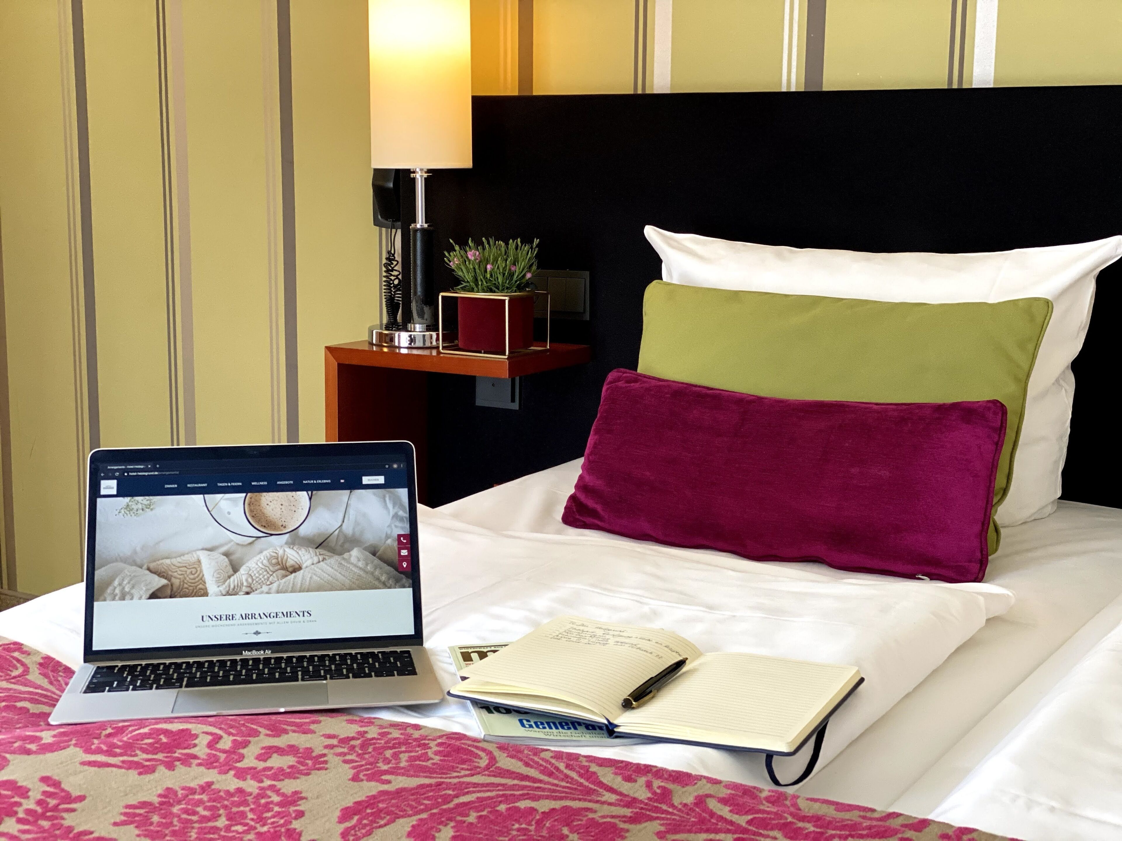 A hotel room with a bed, a laptop, a notebook, and a lamp.