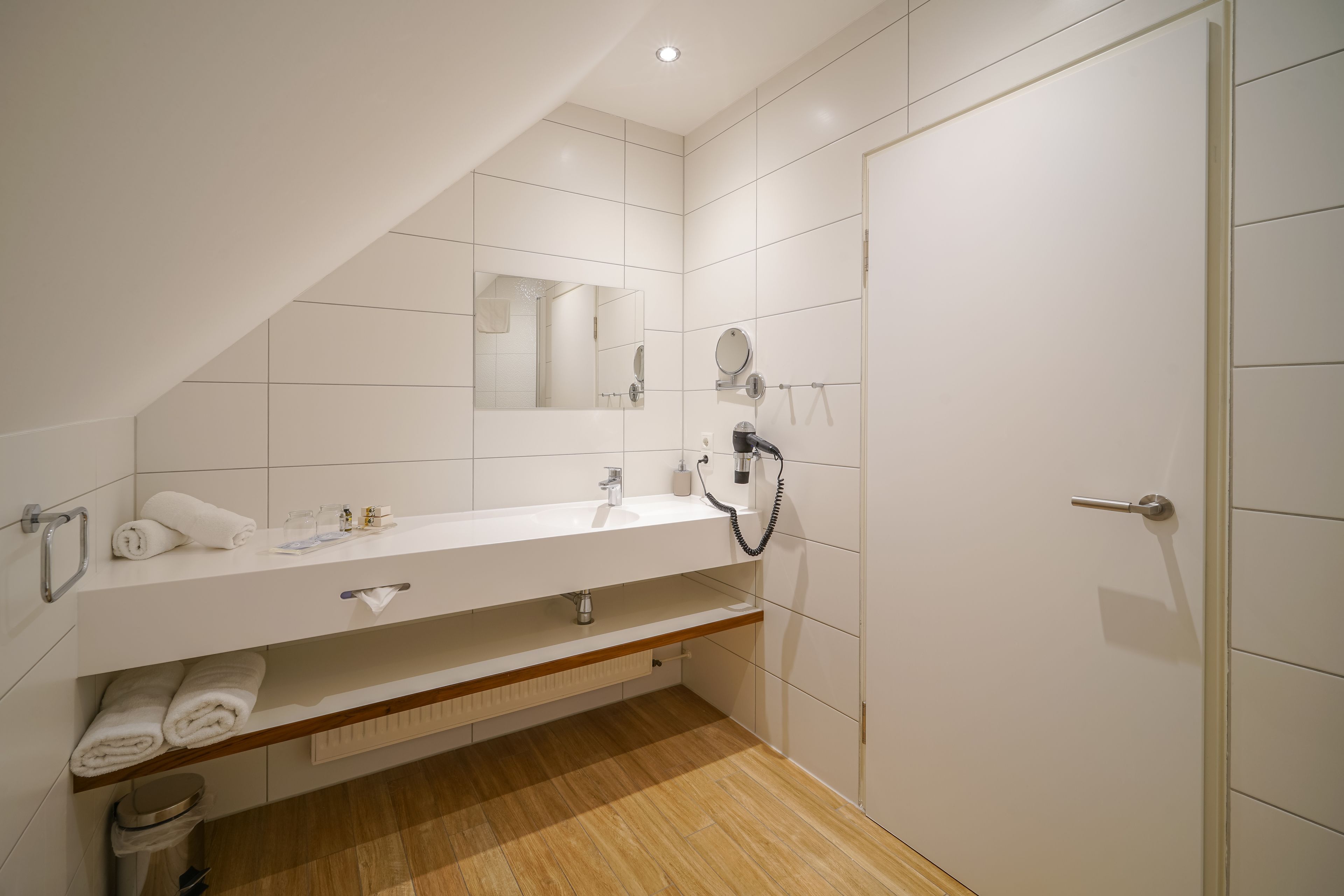 Ein modernes Badezimmer mit weißen Fliesen, Holzboden, Waschtisch inklusive Spiegel und einer Dusche.