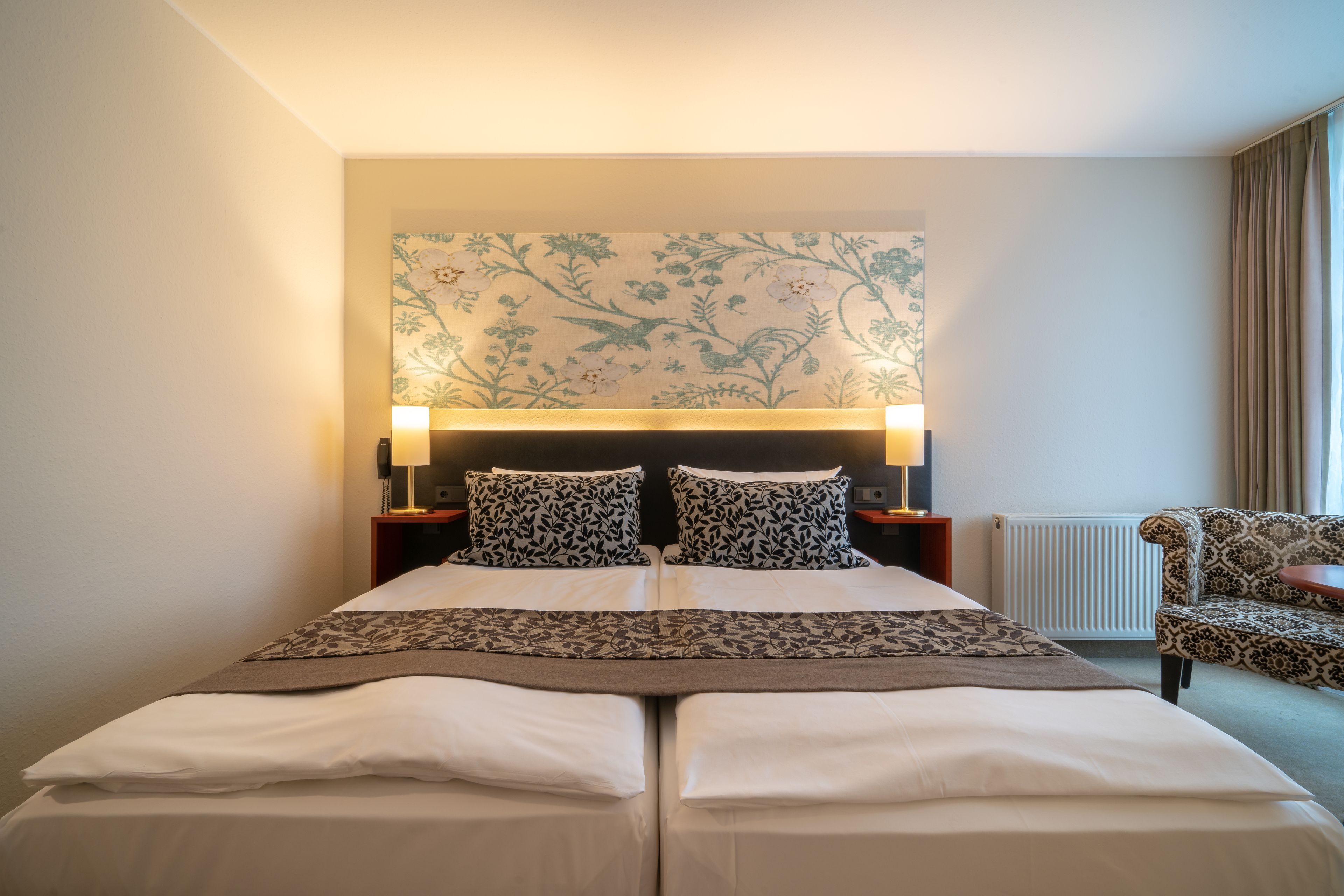 A double bed with a floral headboard, two bedside tables with lamps, a patterned armchair, and a radiator.