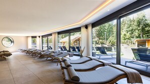 A spa room with sun loungers aligned along a wall with large windows, a round mirror, and a view of an outdoor area with lounge chairs. - Heidegrund (Garrel)