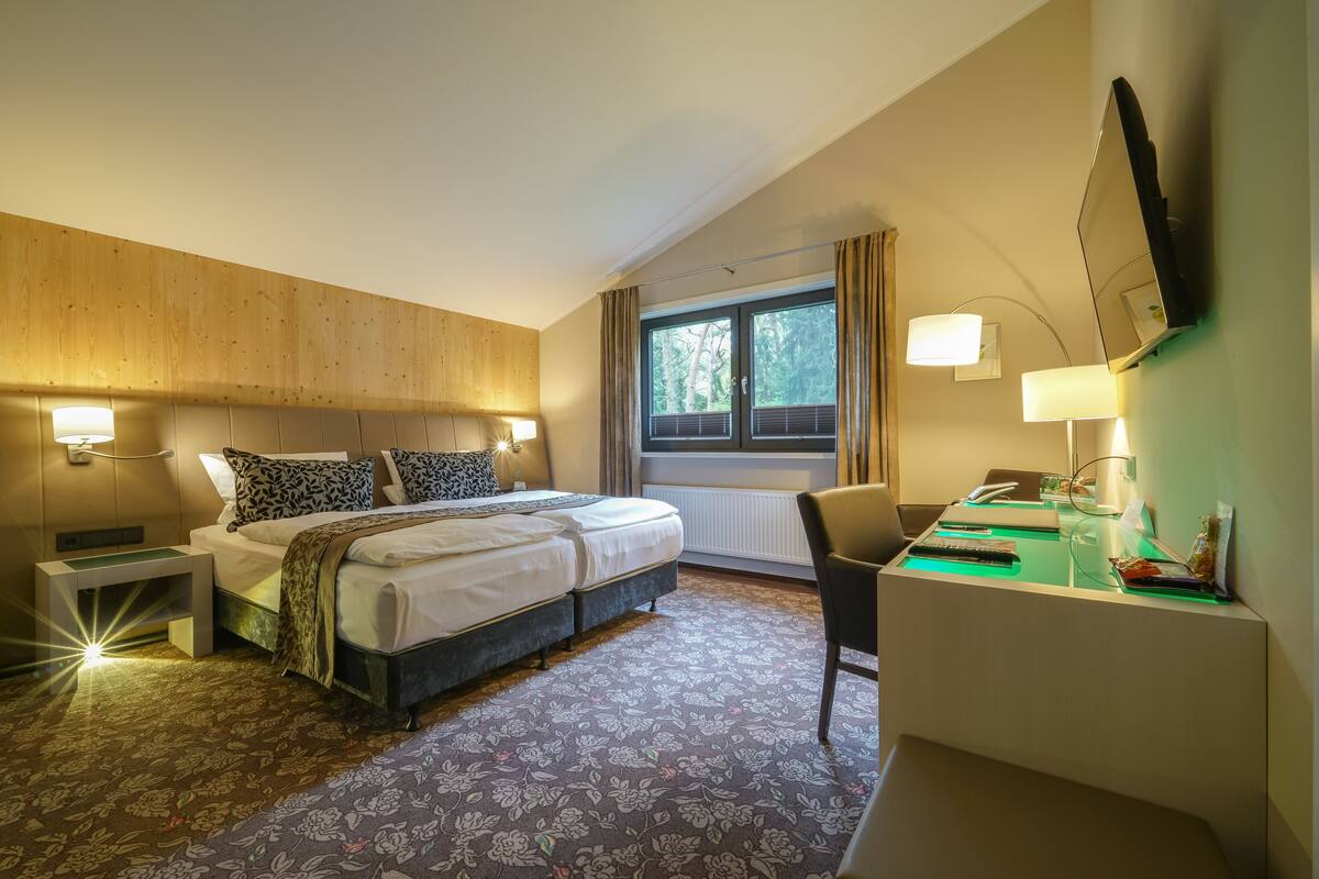 Ett hotellrum med en säng, sängbord, ett skrivbord, en stol, ett fönster med gardiner och en väggmonterad TV.