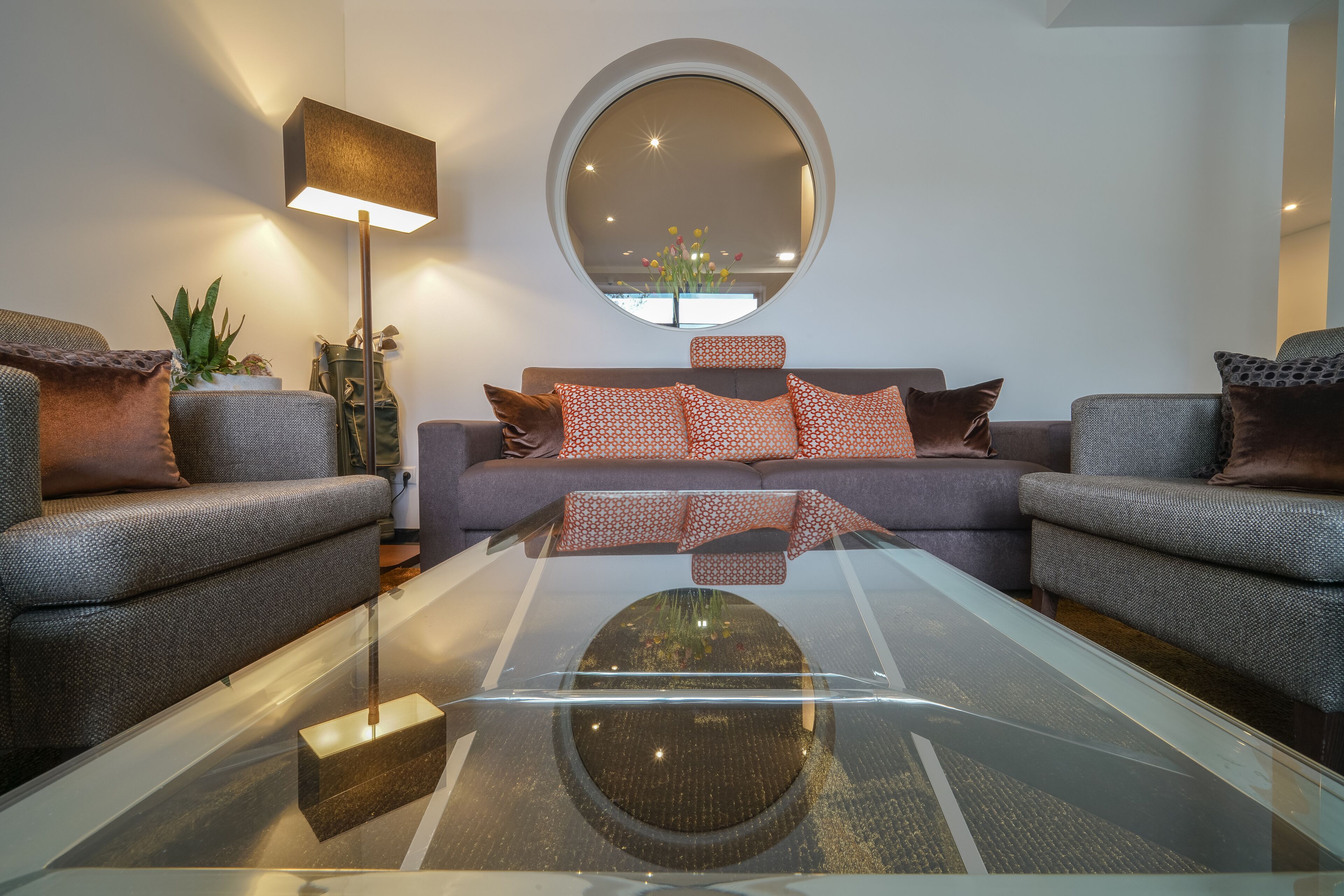 A modern living room with a glass coffee table, grey sofas, patterned cushions, a round mirror, and a floor lamp.