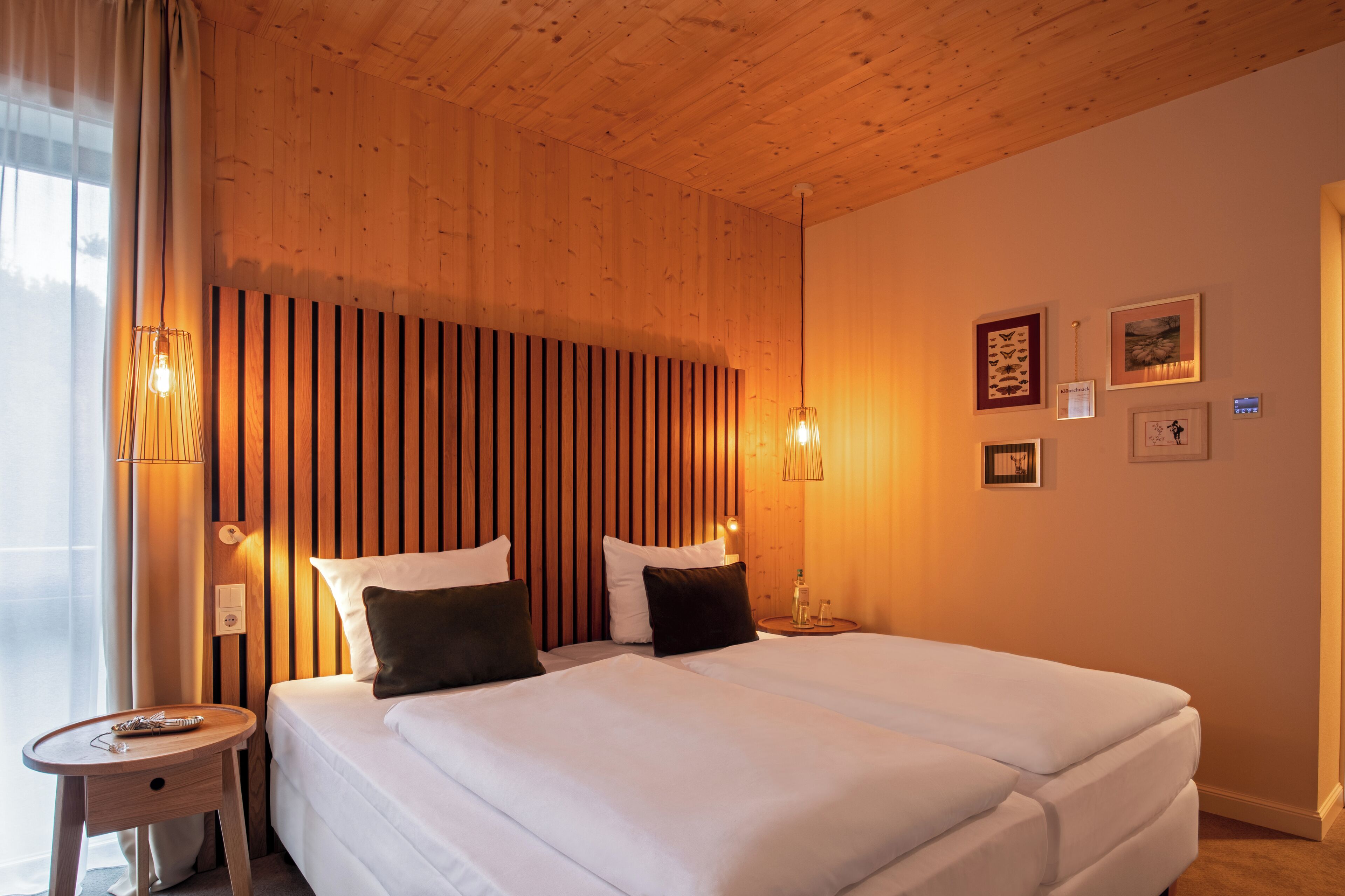 A hotel room with a wooden ceiling, two beds with white linens, a bedside table with a lamp, and wall-mounted artwork.