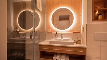 A modern bathroom with a glass shower enclosure, a wooden vanity with a large mirror, and a built-in shelf with towels.
