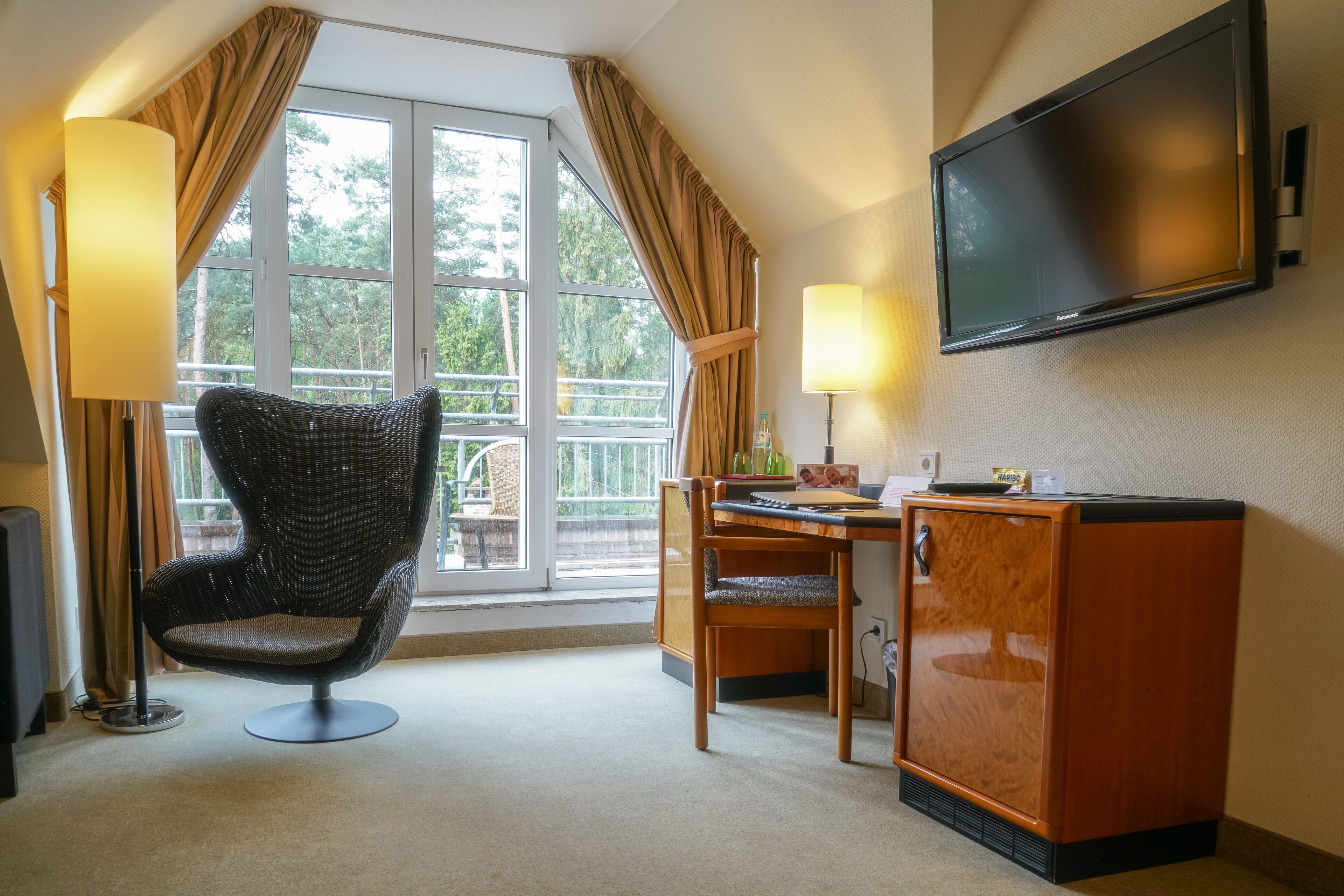 A hotel room with a large window, a wicker chair, a wooden dining table, a television, and a minibar.