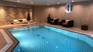 A modern indoor swimming pool with a tiled floor, lounge chairs, and a stone wall. - Heidegrund (Garrel)