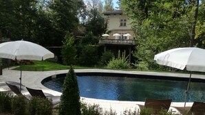 Una piscina con sillones de descanso y sombrillas, rodeada de exuberante vegetación y un edificio con terraza.
