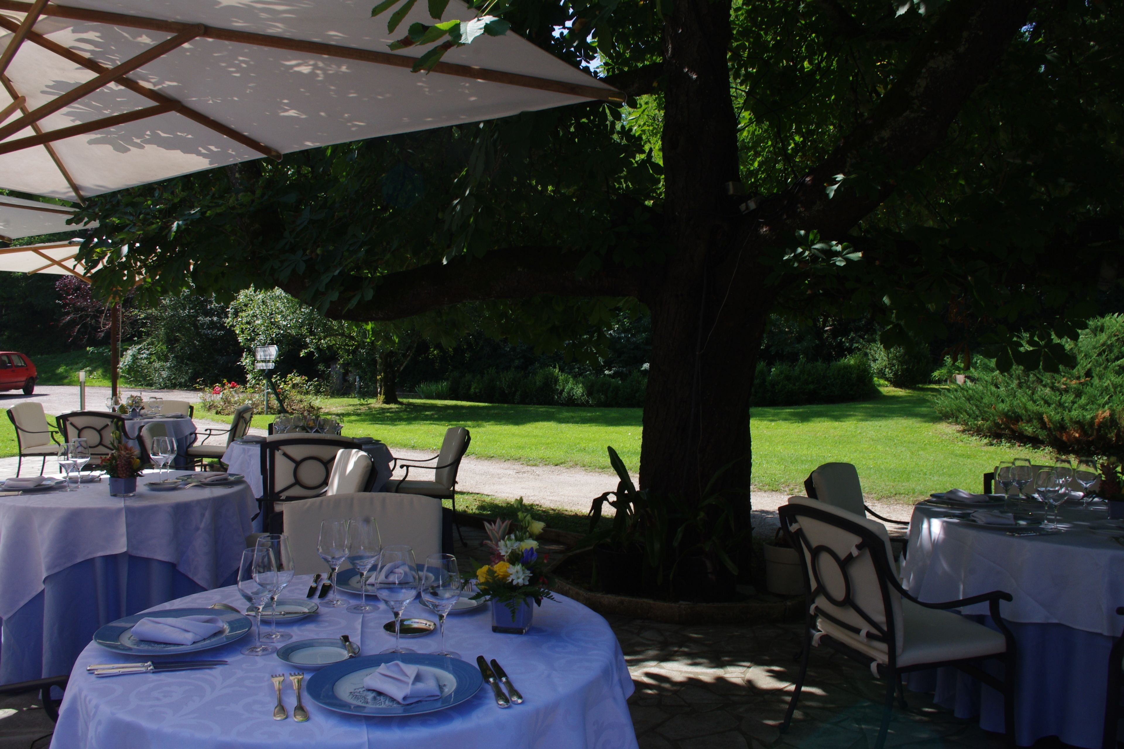 Un área de comedor al aire libre con mesas redondas cubiertas con manteles blancos, dispuestas con platos, vasos y cubiertos.
