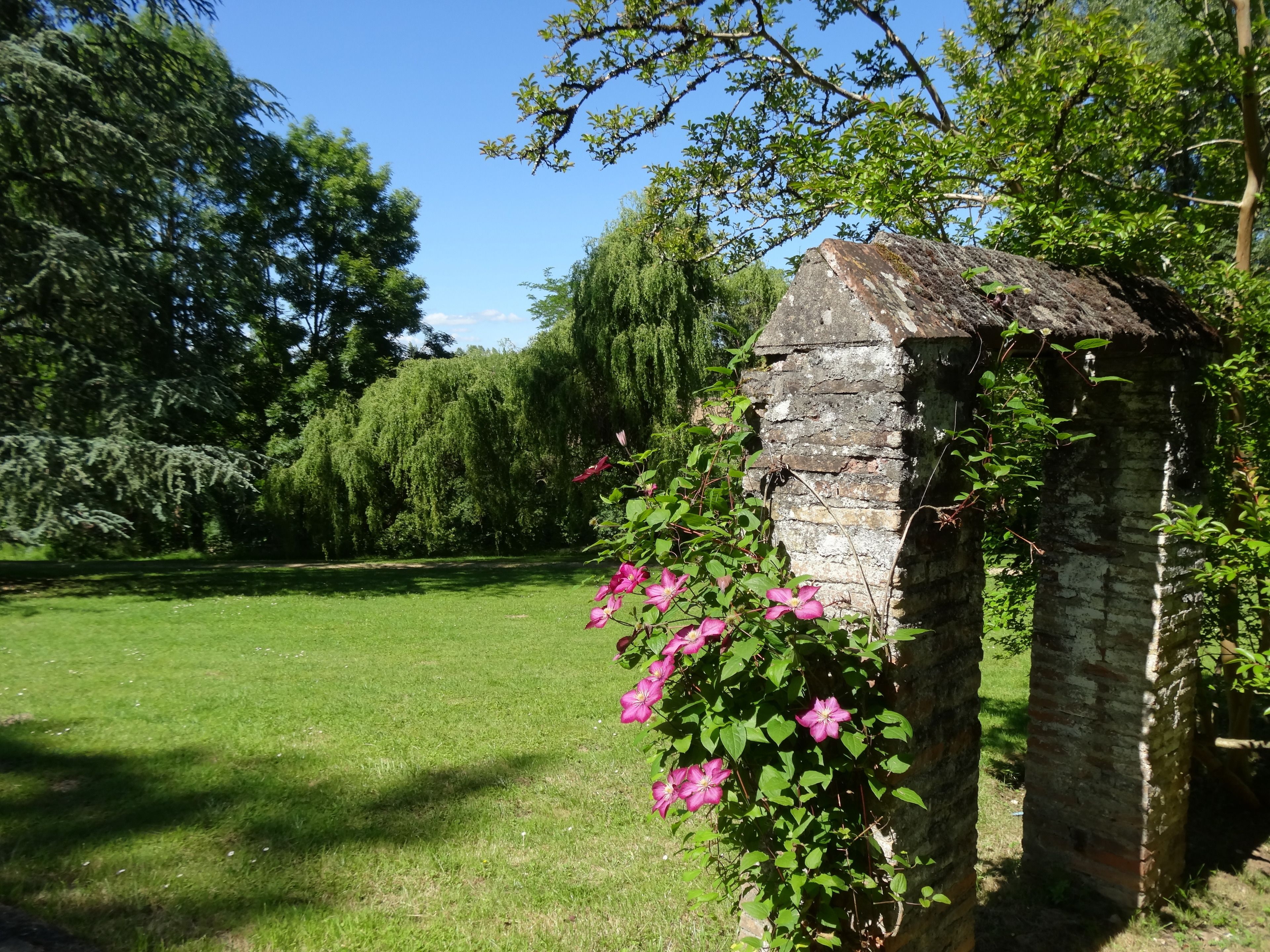 Een stenen gebouw overwoekerd met klimplanten en bloemen, omgeven door een gazon en bomen.