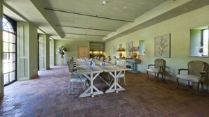 A spacious dining area with a long wooden table and multiple chairs, a kitchen in the background, and large windows on one side.