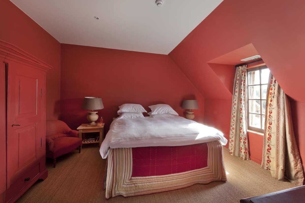 Une chambre avec un plafond en pente, un lit avec une literie blanche, deux lampes de chevet, une porte rouge et une fenêtre ornée de rideaux à motifs floraux.