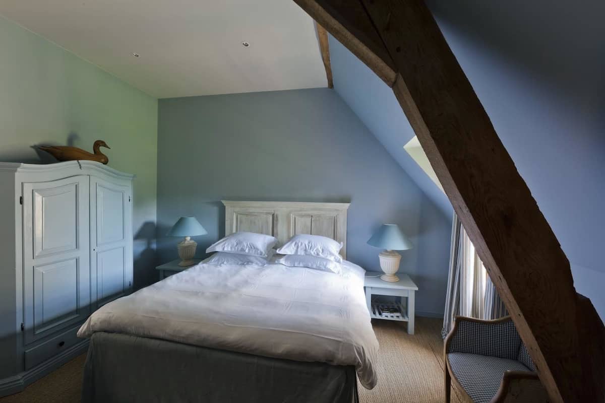 Une chambre avec un plafond en pente, un lit recouvert de draps blancs, deux tables de chevet avec des lampes et une armoire en bois.