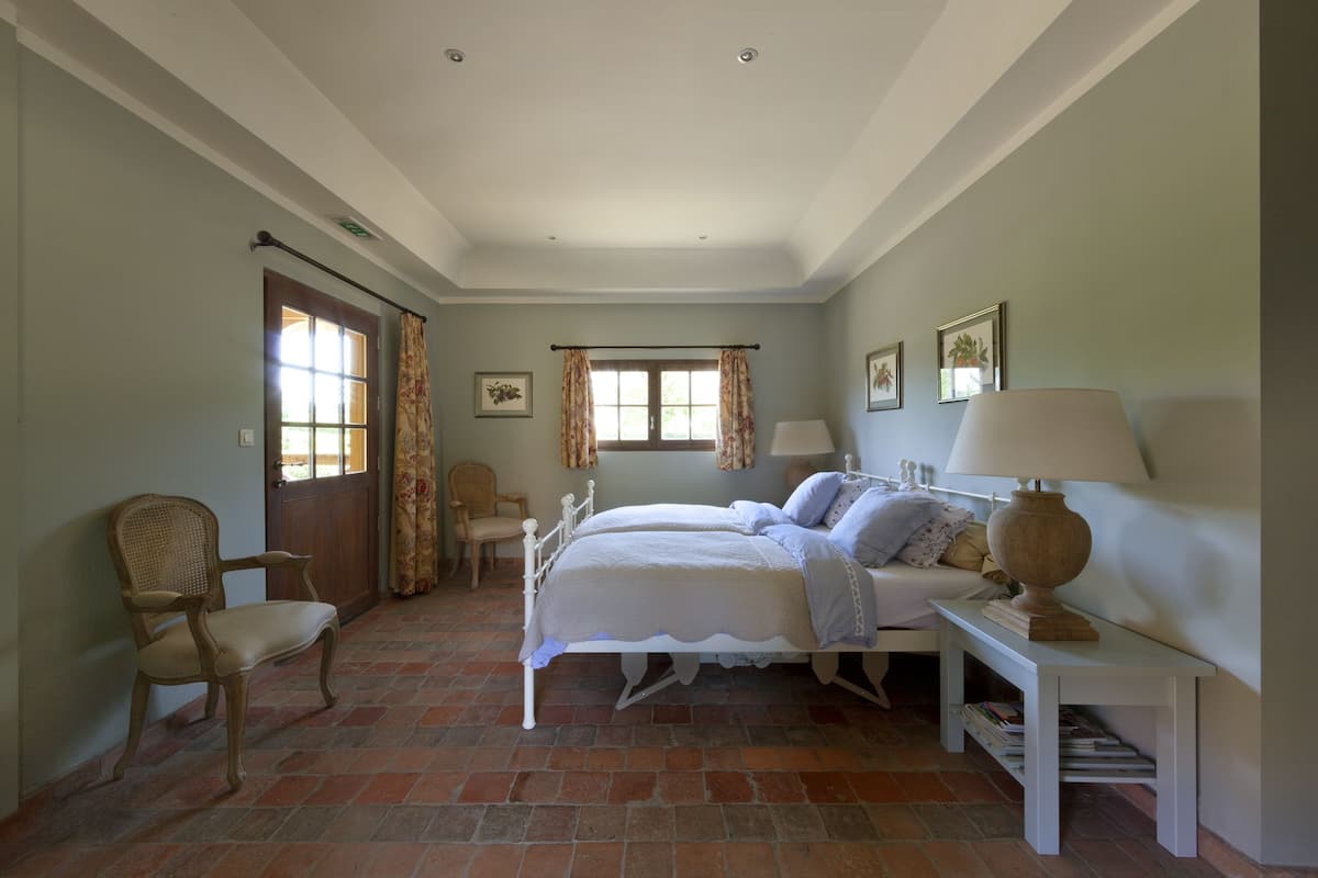 Une chambre à coucher avec un lit double, deux tables de chevet, une chaise, une fenêtre avec des rideaux et une porte.