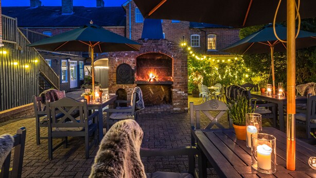 Un patio al aire libre con chimenea de ladrillo, mesas y sillas de madera, rodeado de luces colgantes y sombrillas.