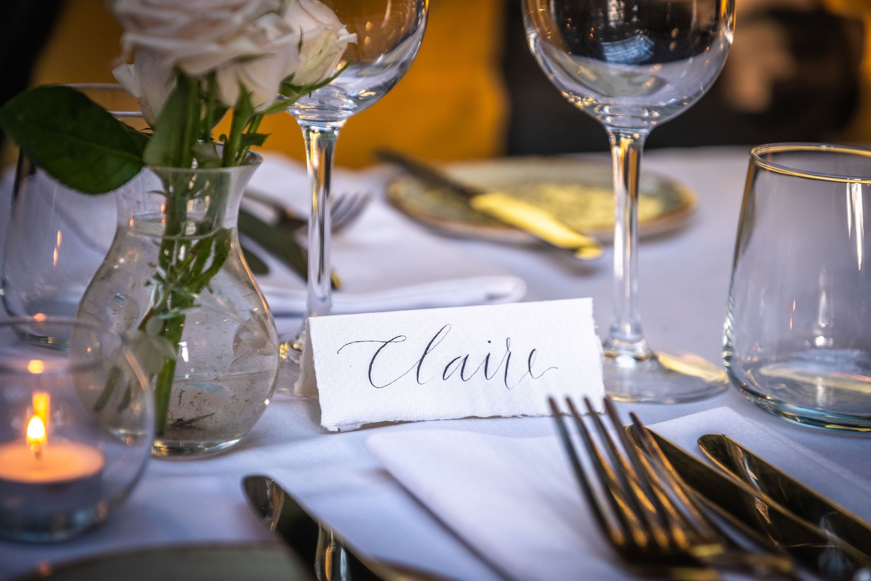 A table set with a name card, a glass vase with a white rose, and a lit candle.