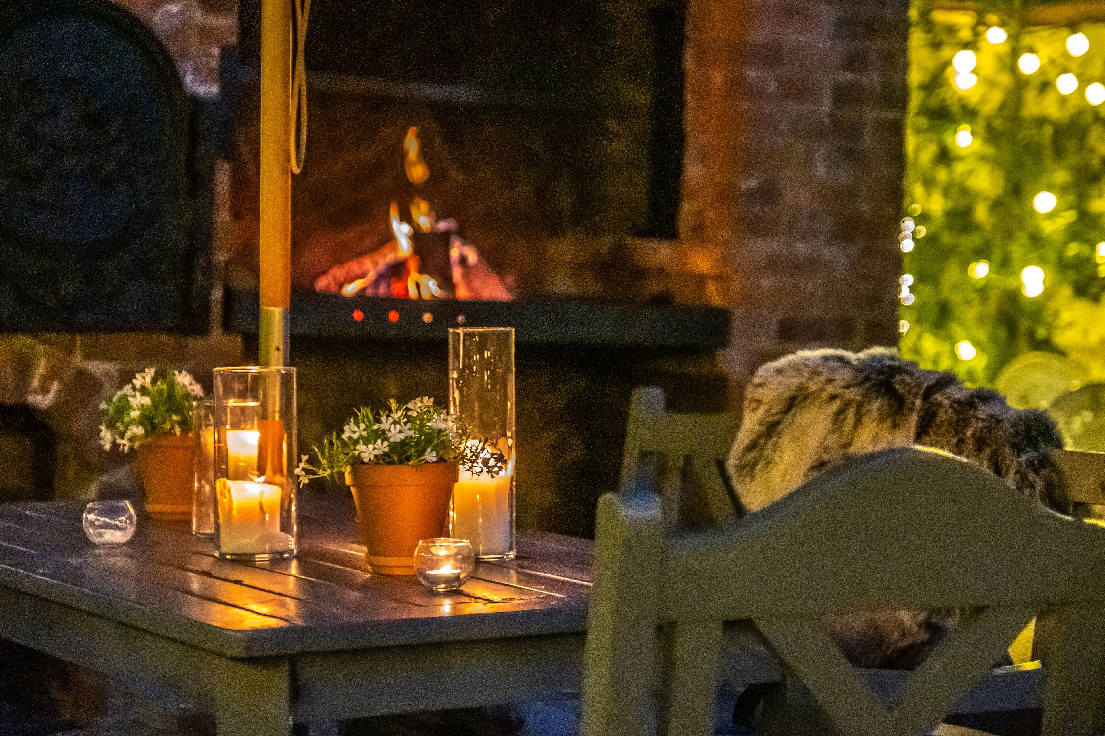 A cozy outdoor seating area with a table set for two, featuring candles, potted plants, and a view of a fireplace.