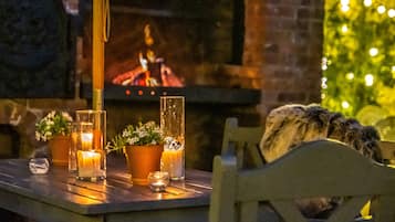A cozy outdoor seating area with a table set for two, featuring candles, potted plants, and a view of a fireplace.