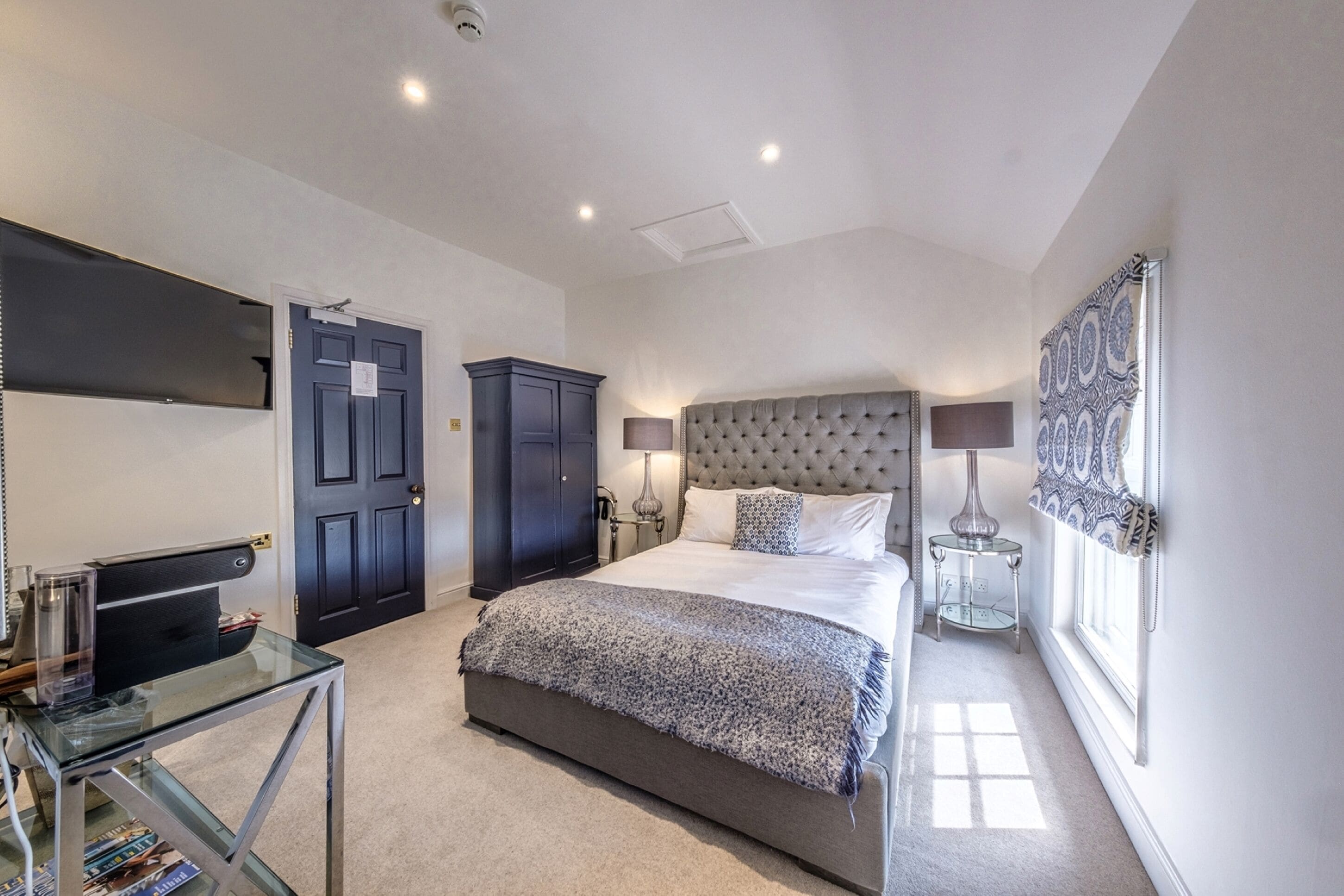 A bedroom with a large bed, a dark wardrobe, a glass table with books, and a lamp.