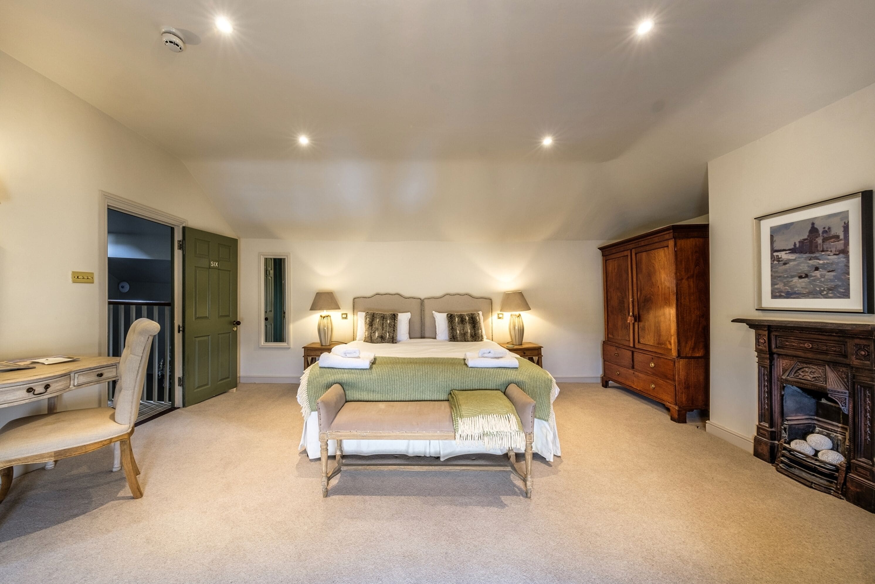 A bedroom with a bed, bedside tables, a desk, a chair, a bench, a wardrobe, and a fireplace.